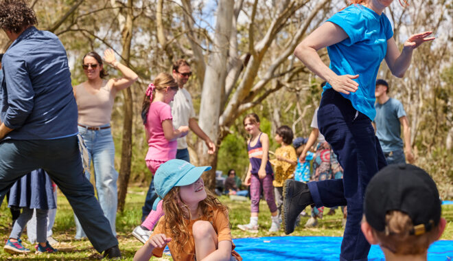 Forest production photo: Laura May Grogan. Polyglot artists dance in a bushland clearing. Children and families around them watch, or join in.