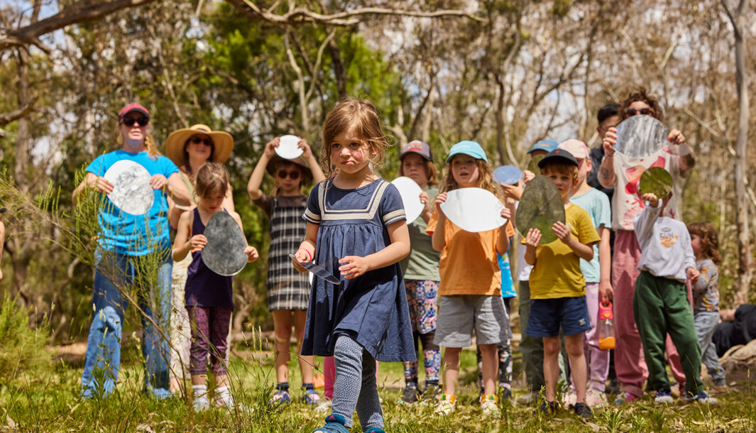 Forest production photo: Laura May Grogan. A child walks determinedly through a bushland clearing. Behind them, a group of children adults and Polyglot artists hold small oblong-shaped mirrors, reflecting light. They are surrounded by trees.