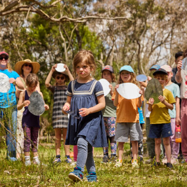 Forest production photo: Laura May Grogan. A child walks determinedly through a bushland clearing. Behind them, a group of children adults and Polyglot artists hold small oblong-shaped mirrors, reflecting light. They are surrounded by trees.