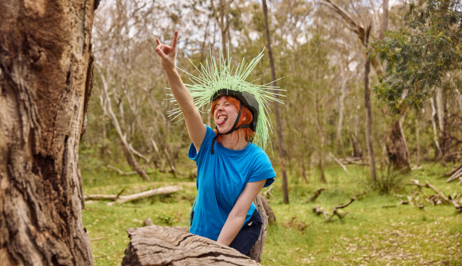 Forest production photo: Laura May Grogan. A Polyglot artist wearing a helmet embellished with long green spikes crouches on a fallen tree trunk, their arm in the air and their tongue out.