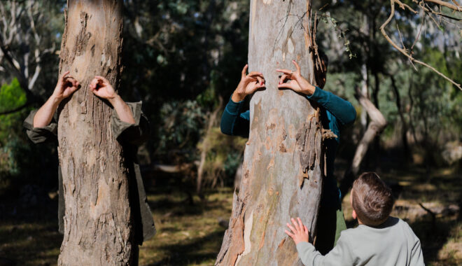 Forest production photo: Sarah Walker. Polyglot artists stand behind trees, using their hands to create eyes. A child stands next to the tree, looking on. They are in a bushland clearing.