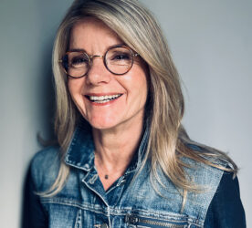 A portrait of Ally Harvey. She has blonde hair, and wears round glasses and a denim jacket. She is in front of a grey background, smiling off-camera.