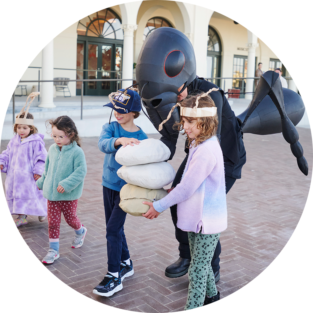 Ants production photo: Lucy Parakhina, Bondi Festival. Children in handmade paper antennae help a Polyglot artist in an intricate black Ant costume carry large breadcrumbs. 