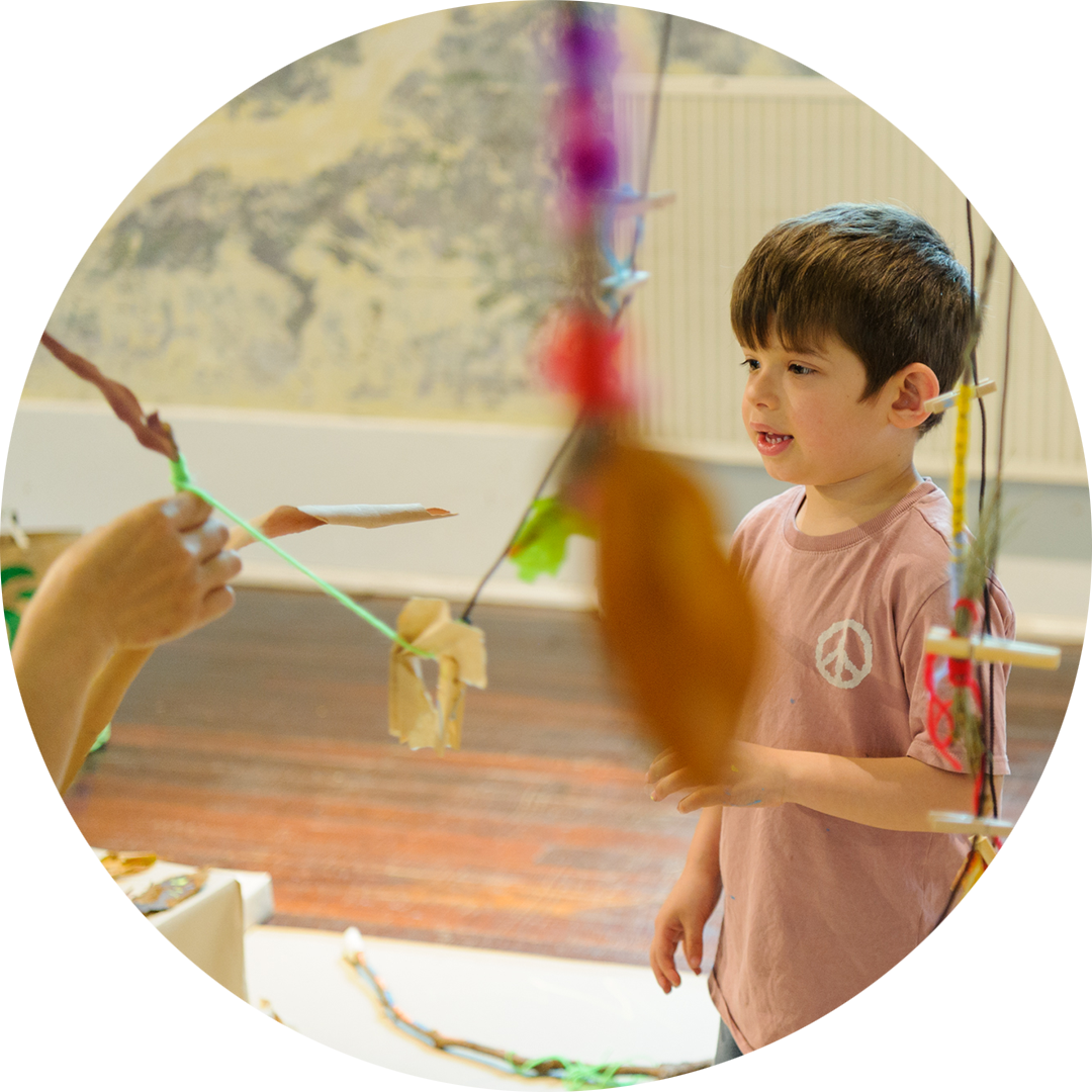 Art Tree production photo: Sarah Walker. A child creates and plays with a Polyglot artist (only their hands are visible). They use leaves and green yarn. There are dangling leaves and colourful creations in front of the lens. 