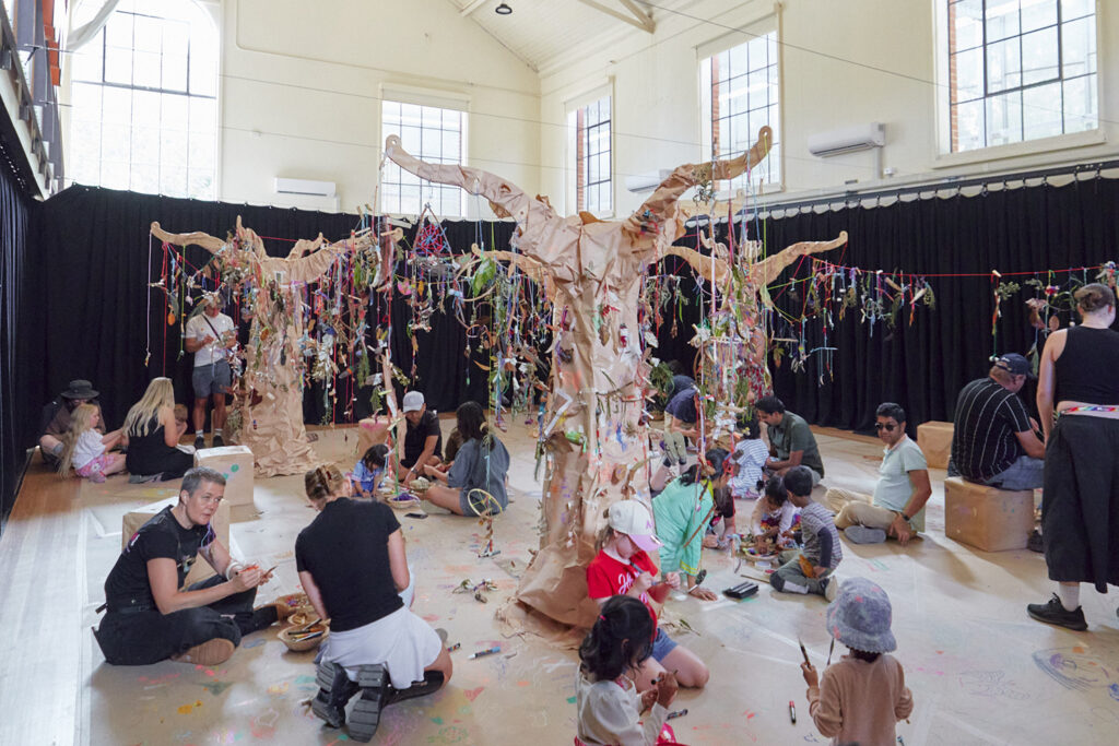 Art Tree production photo: Instant Crush, Moomba Festival (ArtPlay). Wooden trees wrapped in brown paper grow in a large, naturally lit room. Creations made from natural materials hang from the trees with colourful yarn. Children, families and Polyglot artists are gathered on the floor, creating and playing. 