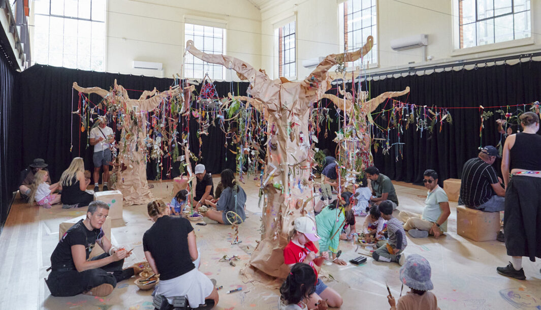 Art Tree production photo: Instant Crush, Moomba Festival (ArtPlay). Wooden trees wrapped in brown paper grow in a large, naturally lit room. Creations made from natural materials hang from the trees with colourful yarn. Children, families and Polyglot artists are gathered on the floor, creating and playing.