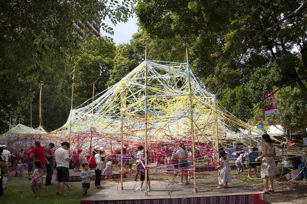 Tangle production photo: Wendy Kimpton. Under a canopy of trees, hundreds of strands of colourful elastic are woven in and around tall golden poles. Children and families create and play among the installation.