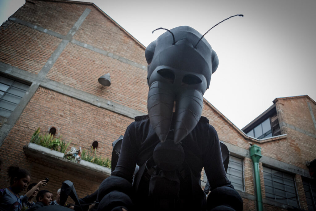 Ants production photo: Matheus Jose Maria. A Polyglot artist in an intricate black Ant costume looks down at the camera. They are in front of a large brick building, with children visible in the background. 