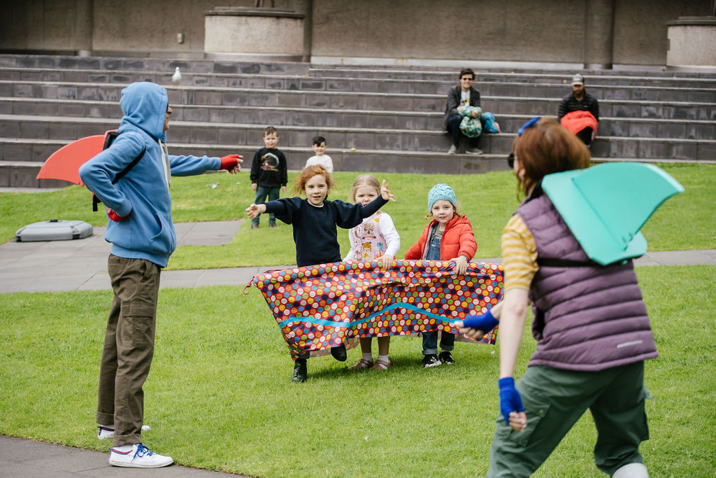 Boats production photo: Jason Lau, Arts Centre Melbourne. Three small children hold a colourful vessel around themselves, engaging with two Polyglot artists in colourful shark fins. They are on a green lawn, with other children and adults looking on. 