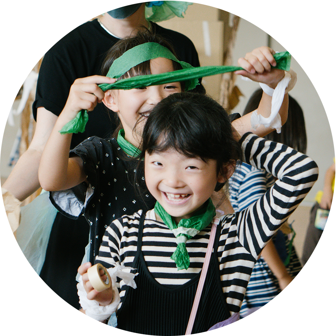 Paper Planet production photo: Ai Ueda. A child in a stripy black and white t-shirt smiles at the camera. They have tape in their hand and are holding their hair up. The child behind them wears a green paper bandana and is in the process of tying green paper onto the other child.