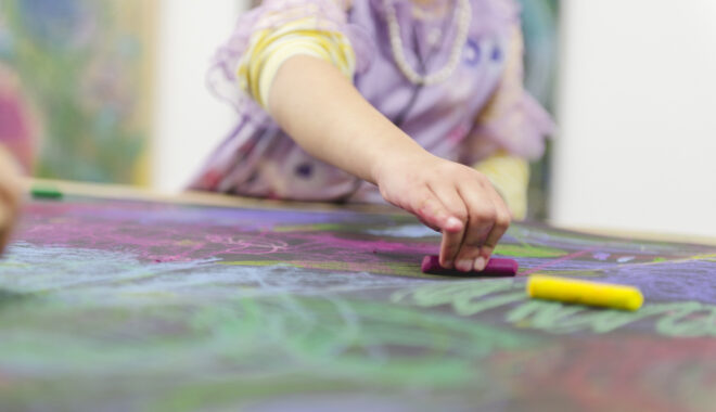 Sound of Drawing production photo: Theresa Harrison. A child in a lilac dress draws on dark paper with soft coloured pastels.