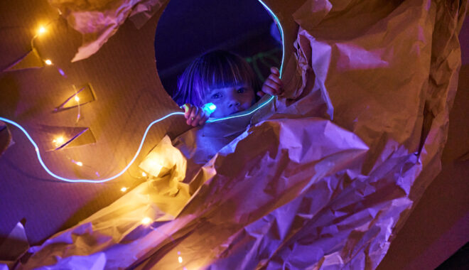 Catch the Light production photo: Laura May Grogan, Flow Festival Australia. A small child with a fringe looks at the camera through a round cardboard window surrounded by brown paper. They are in a darkened space, illuminated only with small string lights.