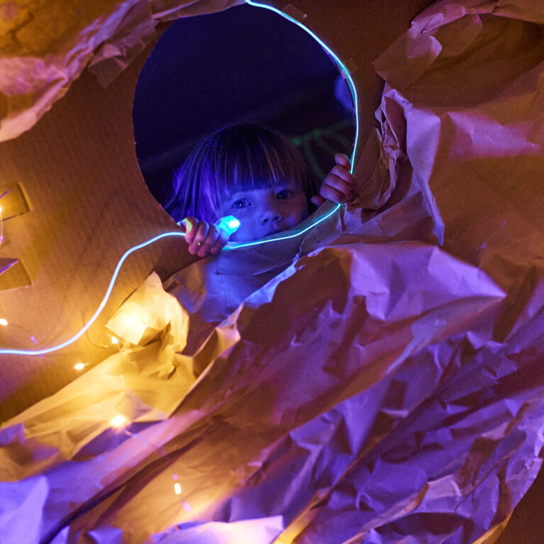 Catch the Light production photo: Laura May Grogan, Flow Festival Australia. A small child with a fringe looks at the camera through a round cardboard window surrounded by brown paper. They are in a darkened space, illuminated only with small string lights.