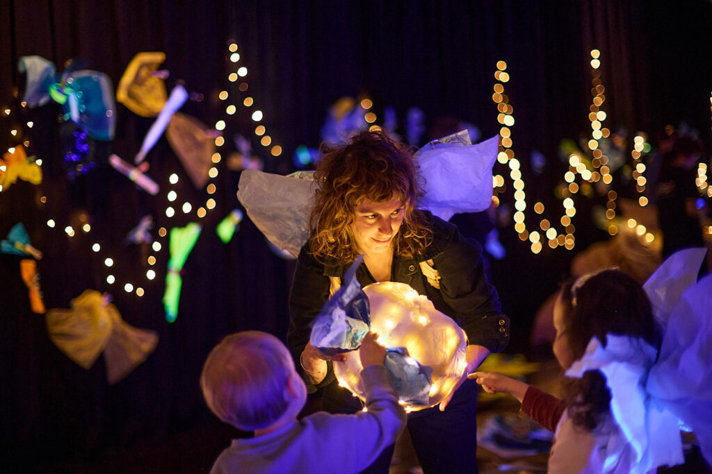 Catch the Light production photo: Laura May Grogan. Two small children, one wearing handmade paper wings, engages with a Polyglot artist who also wears handmade paper wings. They are in a darkened space, illuminated by small lights in the background, and a glowing paper sphere held by the artist. 