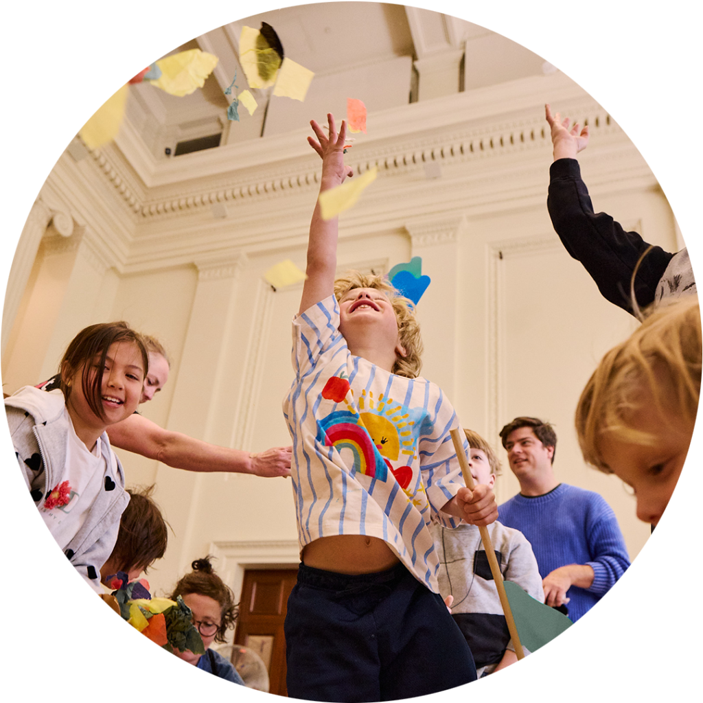 Whirlwind production photo: Laura May Grogan. A child jumps into the air, reaching for swirling colourful paper confetti. Other adults and children reach, jump and move around them.