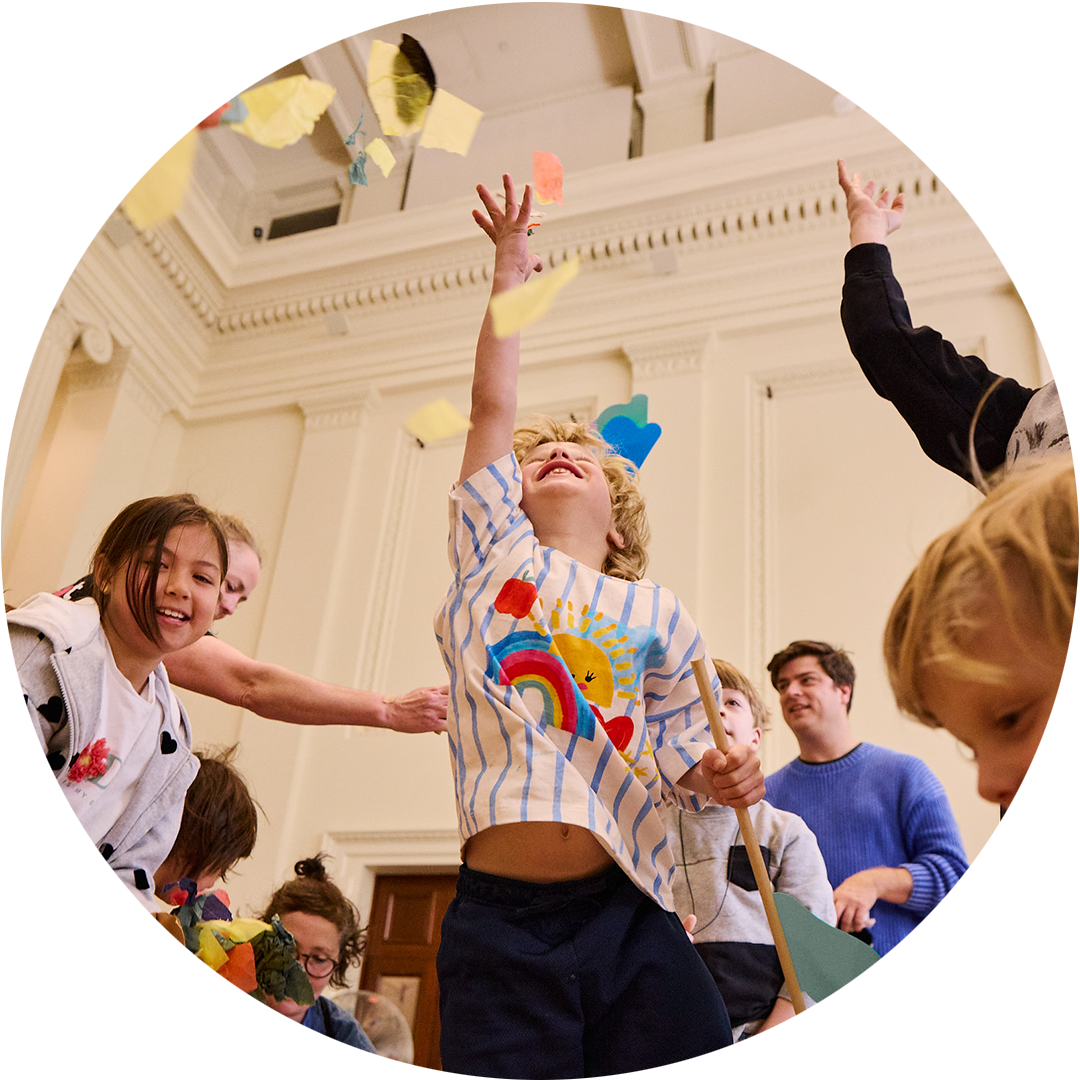 Whirlwind production photo: Laura May Grogan. A child jumps into the air, reaching for swirling colourful paper confetti. Other adults and children reach, jump and move around them.
