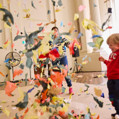 Whirlwind production photo: Laura May Grogan. A child in a red jumper smiles as colourful confetti swirls around them. An adult is in the background, eclipsed by the flying confetti. There is a Polyglot artist to the left of the image.
