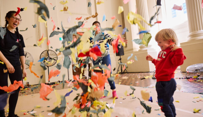 Whirlwind production photo: Laura May Grogan. A child in a red jumper smiles as colourful confetti swirls around them. An adult is in the background, eclipsed by the flying confetti. There is a Polyglot artist to the left of the image.
