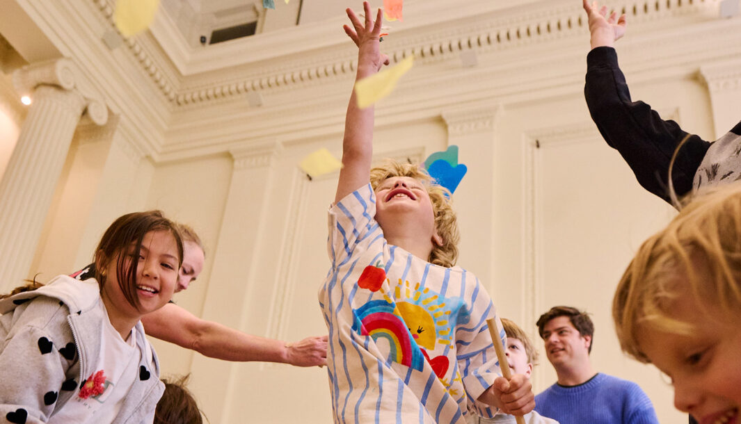 Whirlwind production photo: Laura May Grogan. A child jumps into the air, reaching for swirling colourful paper confetti. Other adults and children reach, jump and move around them.