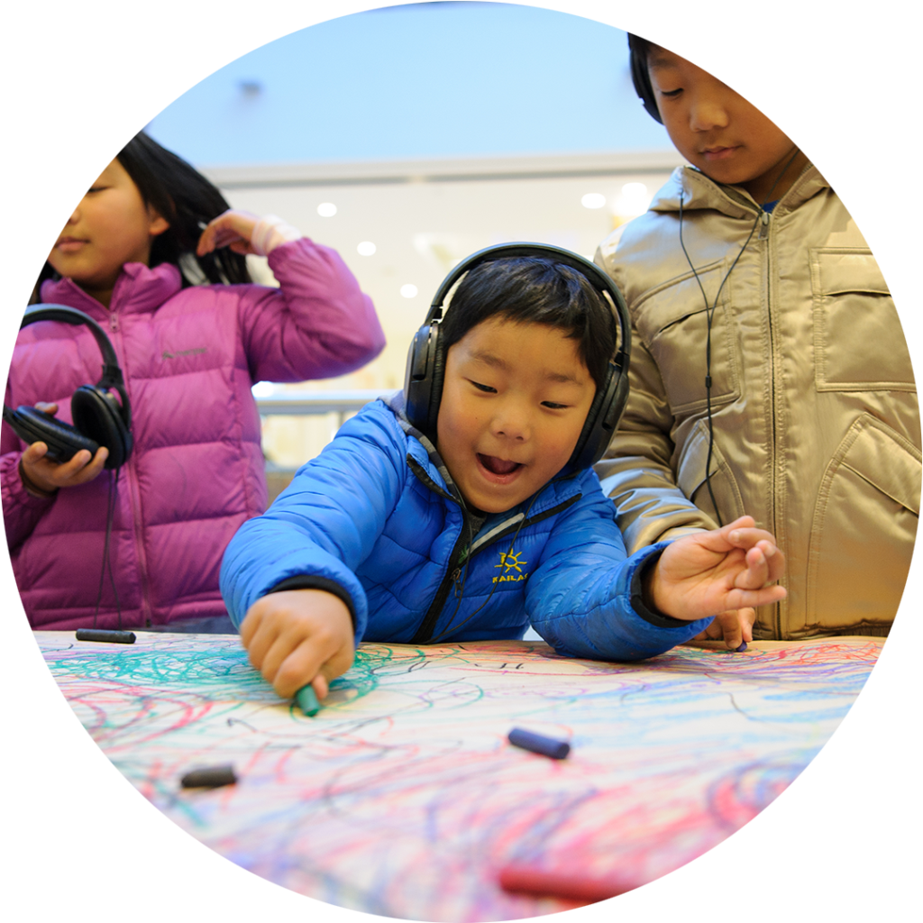 Sound of Drawing production photo. Sarah Walker. Two children wearing headphones stand at a table covered in brown paper, drawing with pastels. A third child stands near them, holding a pair of headphones.