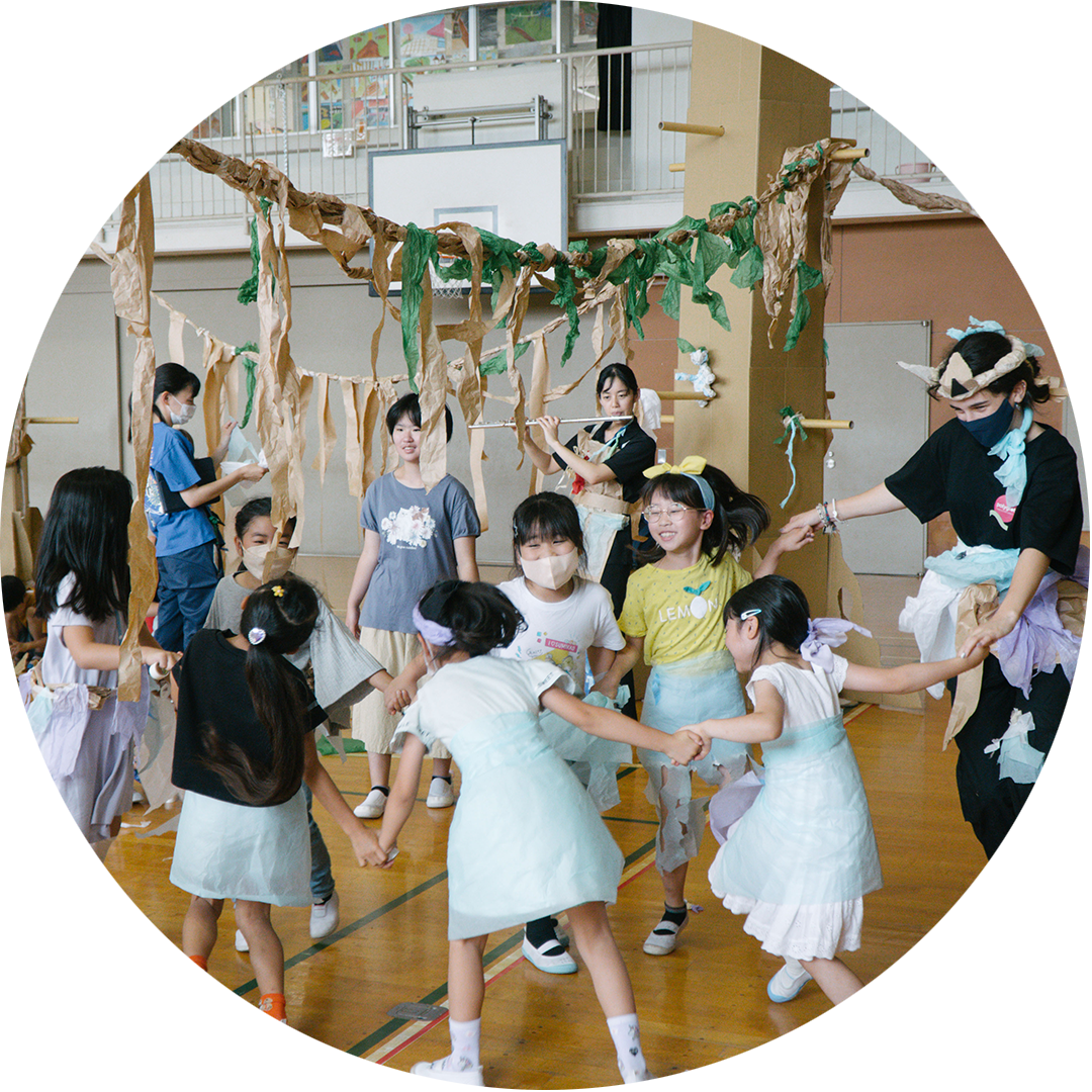 Paper Planet production photo: A group of children hold hands with each other and a Polyglot artist in a circle formation. There is another artist in the background playing the flute, There are children in the background against the set of tall cardboard trees and tissue paper vines.