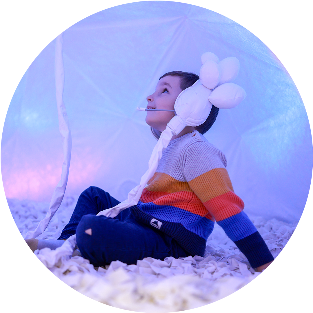 Voice Lab production photo: Pete Wallis. A child in a stripy jumper looks up above. They are wearing intricate white embellished headphones and are seated cross-legged in the Voice Lab space. There is a soft blue lighting wash.