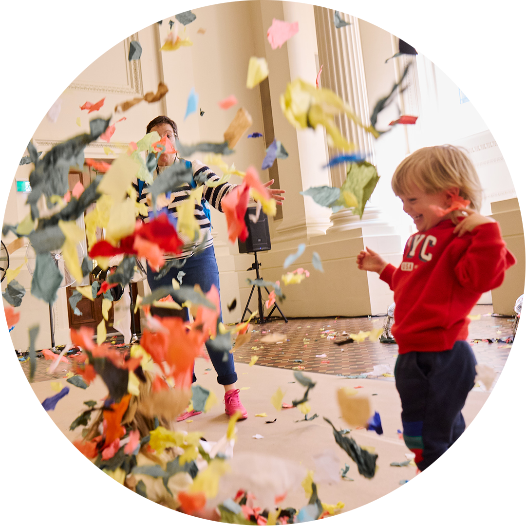 Whirlwind production photo: Laura May Grogan. A child in a red jumper smiles as colourful confetti swirls around them. An adult is in the background, eclipsed by the flying confetti.