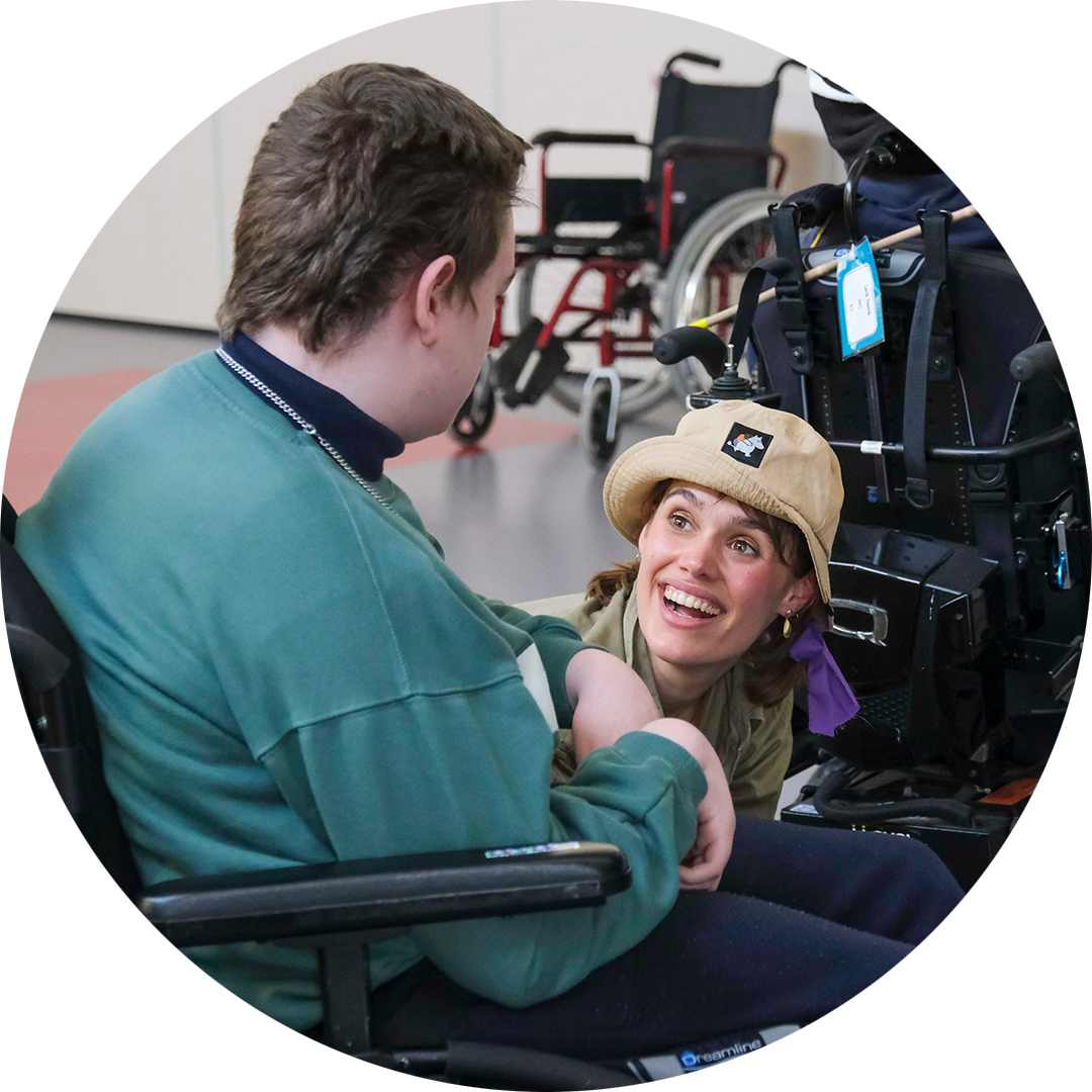 When the World Turns: Suzanne Phoenix. A Polyglot artist crouches towards the ground, wearing a bucket hat and khaki top. They are engaged with a child wearing a teal jumper, who uses a wheelchair.