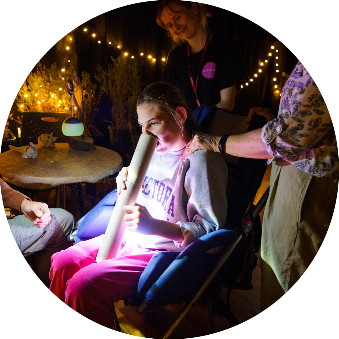 When the World Turns: Sarah Walker. A child who uses a wheelchair, plays with a long cylinder cardboard tube. A Polyglot artist stands behind them, smiling and watching on. Another adult has their hand on the child's shoulder. There are fairy lights in the background.