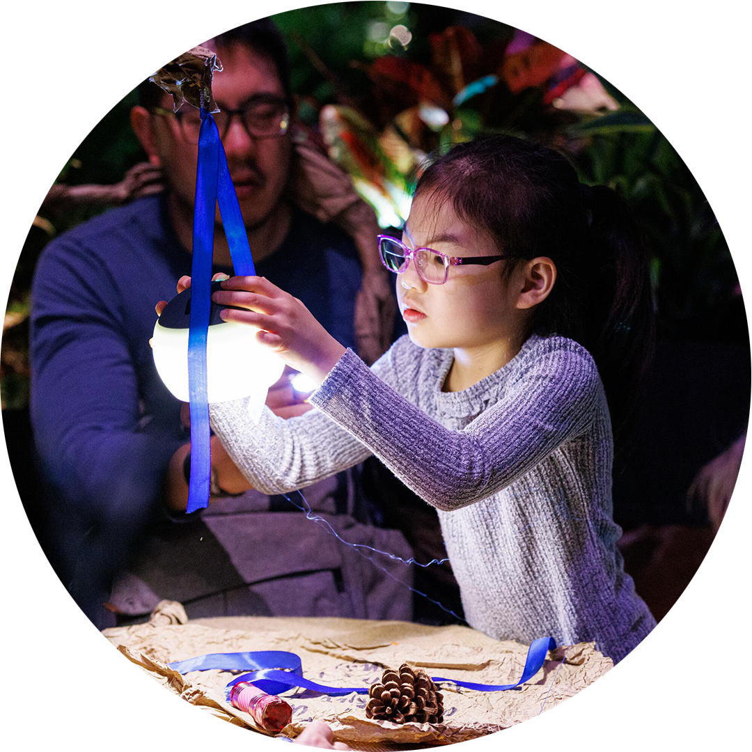 When the World Turns production photo: Sachyn Mital. A child wearing purple glasses reaches toward a light bulb hanging from a blue ribbon. An adult is seated in the background,