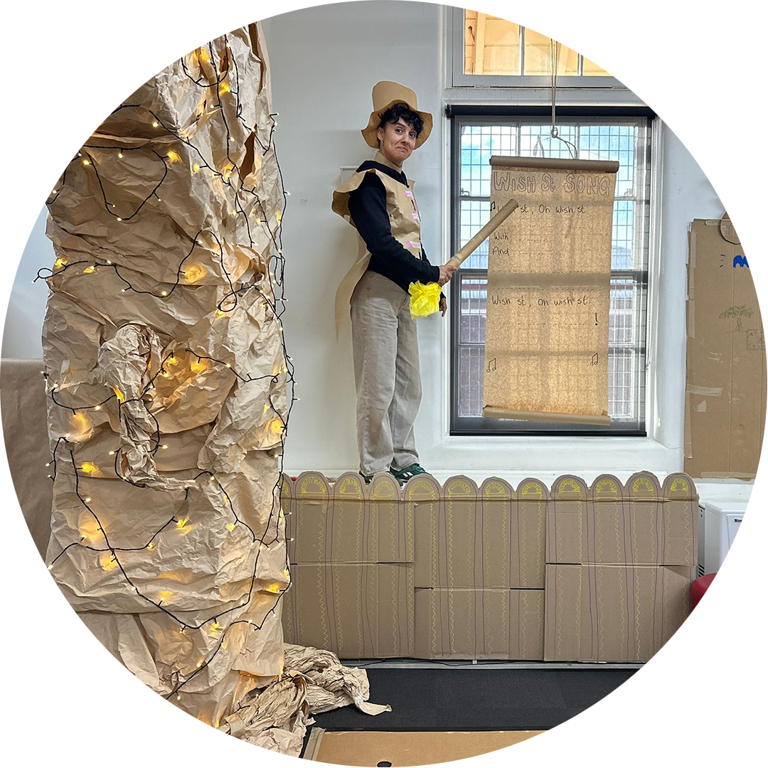 Wish Street production photo: A Polyglot artist stands on a platform in the classroom, gesturing toward a roll of parchment paper with the 'Wish Street song' written on it. They are wearing a paper top hat and vest. On the left side of the image, there's the Wishing Tree trunk with fairy lights wrapped around it..