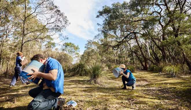 Forest production photo: Laura May Grogan. Polyglot artists dressed in blue crouch in a bushland clearing, studying elements of the landscape through round blue lampshades. A stage manager walks behind them. They are surrounded by trees.