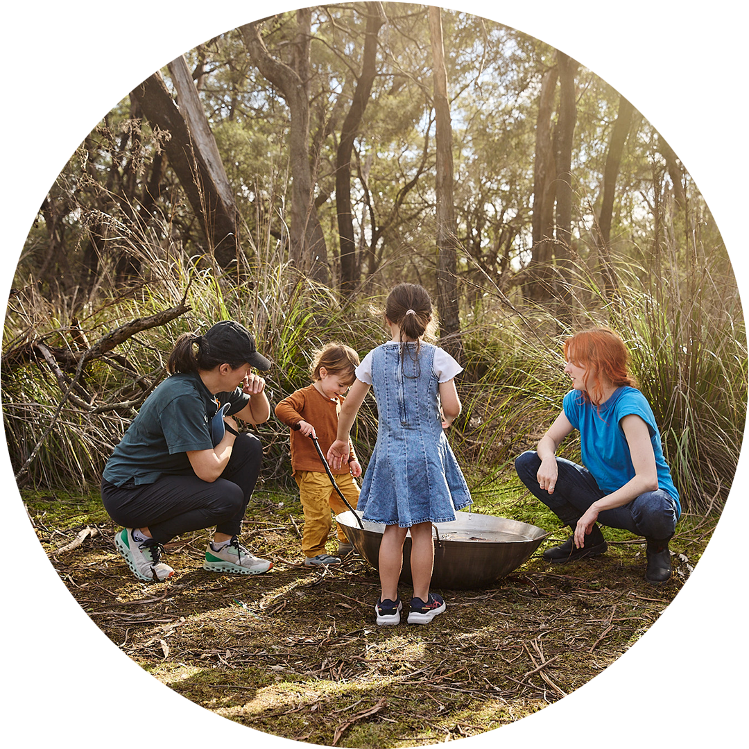 Forest production photo: Laura May Grogan. A Polyglot artist crouches on the forest floor, level with two other children who are standing and playing with long sticks in the silver cauldron of water. An adult is also crouched toward the ground, across the artist.