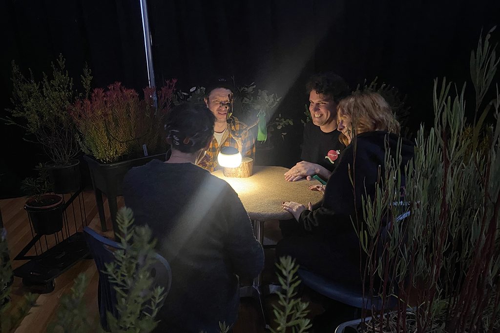 Behind-the-scenes When the World Turns (schools) photo. Four Polyglot artists sit around a small table, illuminated by a lamp. They are surrounded by plants. 