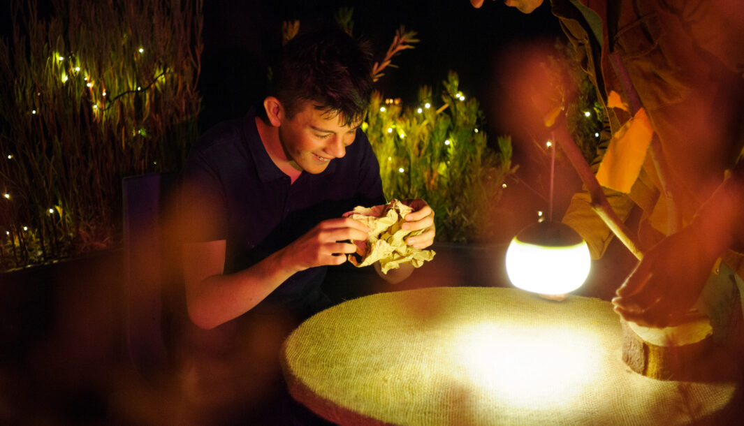 Paper Planet production photo: Sarah Walker. A child and Polyglot artist are smiling and interacting around a round table illuminated by a glowing lantern in a warmly lit outdoor setting with string lights.