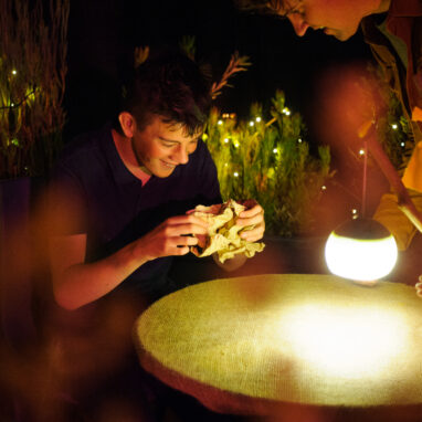 Paper Planet production photo: Sarah Walker. A child and Polyglot artist are smiling and interacting around a round table illuminated by a glowing lantern in a warmly lit outdoor setting with string lights.