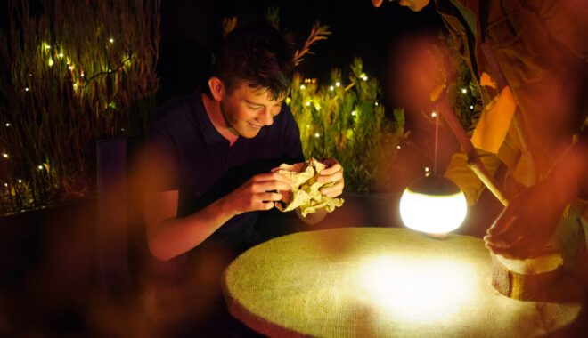 Paper Planet production photo: Sarah Walker. A child and Polyglot artist are smiling and interacting around a round table illuminated by a glowing lantern in a warmly lit outdoor setting with string lights.