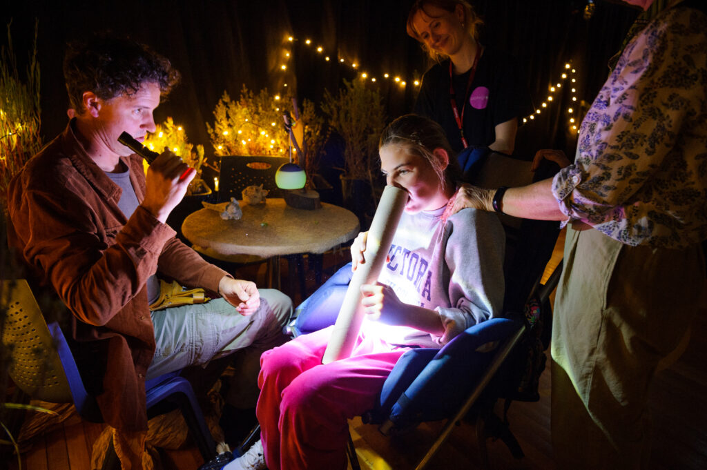 When the World Turns production photo: Sarah Walker. A group of Polyglot artists and educators are gathered in a dimly lit outdoor setting with string lights, where one child is sitting and holding a large white tube.