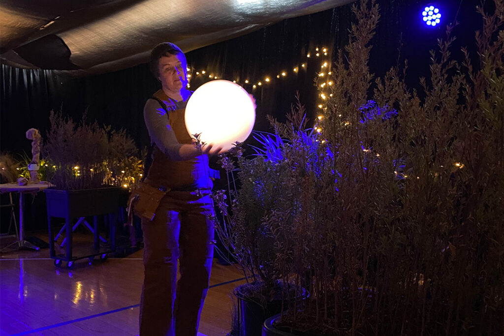 Behind-the-scenes When the World Turns (schools) photo: Maisy Butchart. A Polyglot artist holds an illuminated moon in their hands, holding their gaze towards it. They are standing in an open space filled with plants and warm fairy lights.