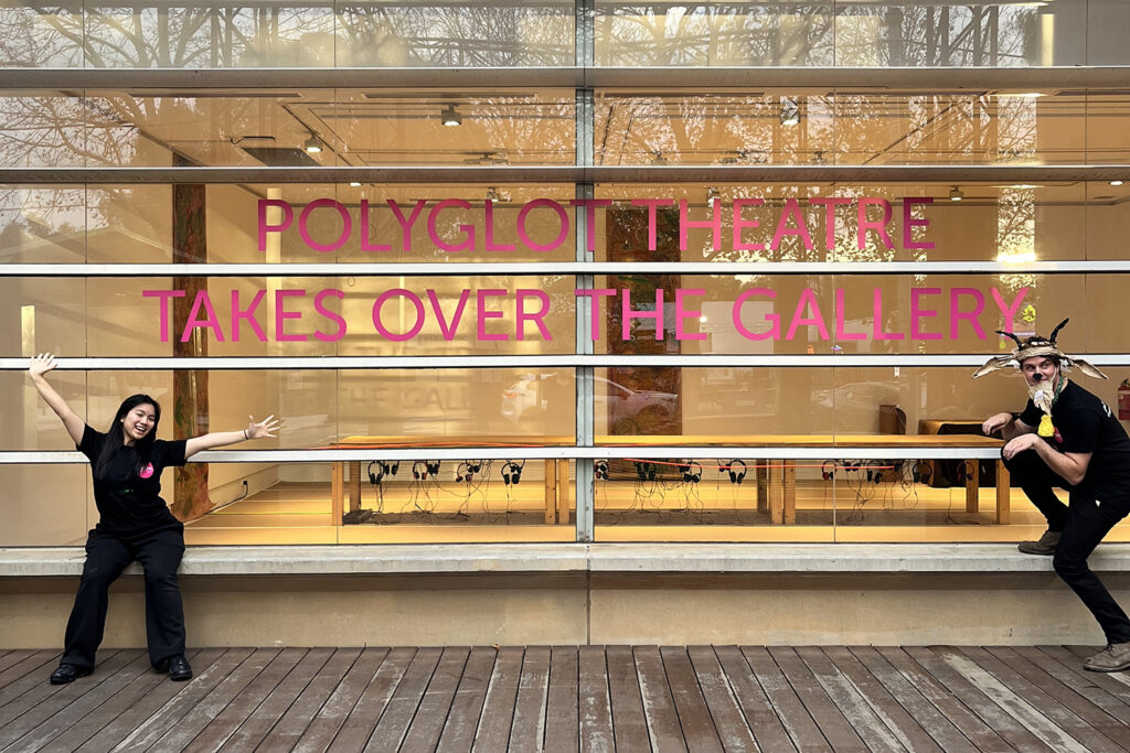 Behind-the-scenes photo: Julie Wright. Two Polyglot artists, one wearing a handmade paper costume, pose either side of a large sign on the front window of Wyndham Art Gallery. It reads, 'Polyglot Theatre Takes Over the Gallery. The Sound of Drawing table is visible inside.