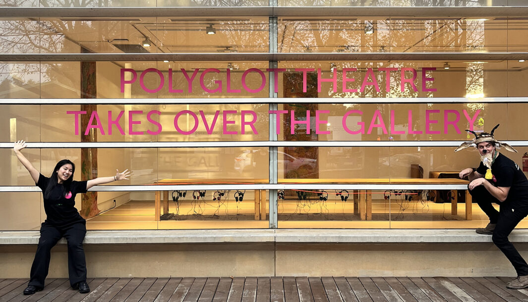 Behind-the-scenes photo: Julie Wright. Two Polyglot artists, one wearing a handmade paper costume, pose either side of a large sign on the front window of Wyndham Art Gallery. It reads, 'Polyglot Theatre Takes Over the Gallery. The Sound of Drawing table is visible inside.