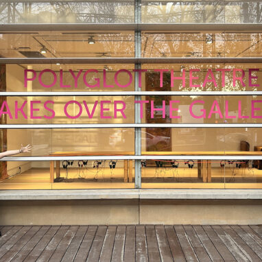 Behind-the-scenes photo: Julie Wright. Two Polyglot artists, one wearing a handmade paper costume, pose either side of a large sign on the front window of Wyndham Art Gallery. It reads, 'Polyglot Theatre Takes Over the Gallery. The Sound of Drawing table is visible inside.