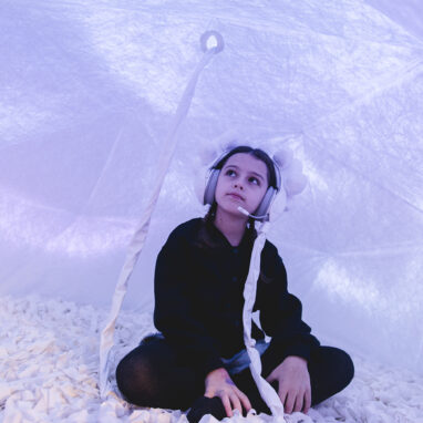 Voice Lab production photo: Theresa Harrison. A child sits cross legged in a soft, white enclosed space, illuminated with lilac light. They wear embellished white headphones connected to the roof of the dome via a white cord.