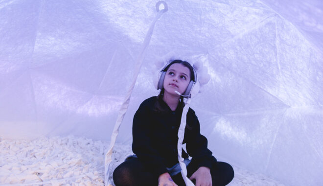 Voice Lab production photo: Theresa Harrison. A child sits cross legged in a soft, white enclosed space, illuminated with lilac light. They wear embellished white headphones connected to the roof of the dome via a white cord.