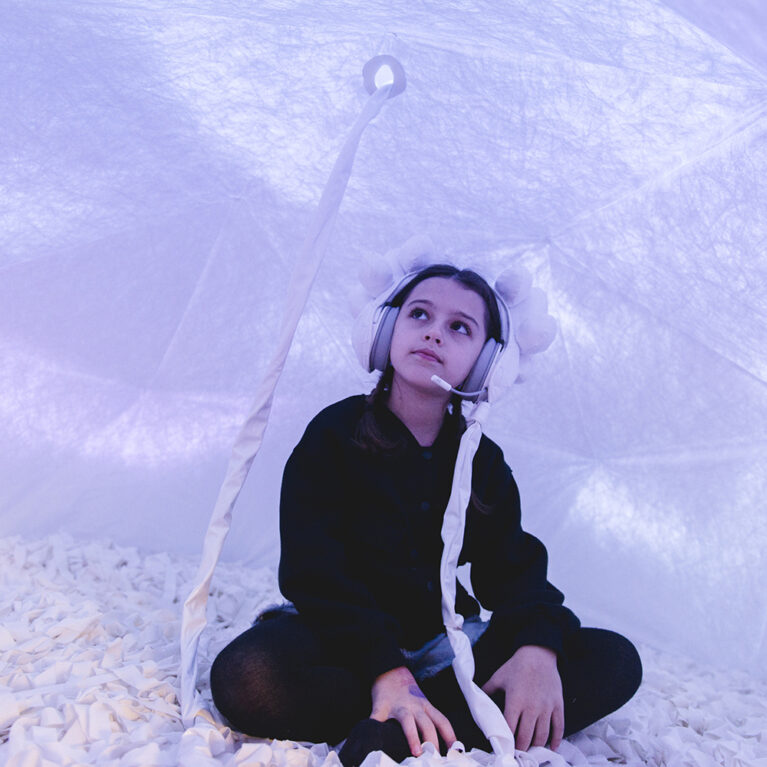 Voice Lab production photo: Theresa Harrison. A child sits cross legged in a soft, white enclosed space, illuminated with lilac light. They wear embellished white headphones connected to the roof of the dome via a white cord.